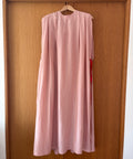 A light pink sleeveless long dress with a high round neckline hangs on a wooden hanger against a brown wooden door.