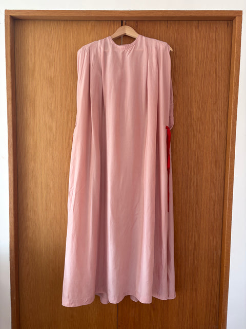 A light pink sleeveless long dress with a high round neckline hangs on a wooden hanger against a brown wooden door.