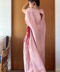 A woman wearing a loose, long pink dress or kaftan with red ribbon ties, standing in front of a wooden door in an indoor setting.