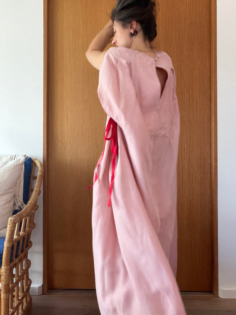 A woman wearing a loose, long pink dress or kaftan with red ribbon ties, standing in front of a wooden door in an indoor setting.