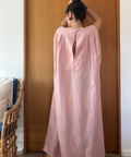 A woman viewed from behind wearing a loose, long dusty pink dress or tunic with a high open back, standing in front of a wooden door in an indoor setting.