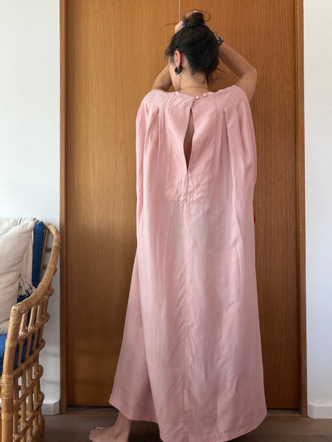 A woman viewed from behind wearing a loose, long dusty pink dress or tunic with a high open back, standing in front of a wooden door in an indoor setting.