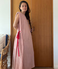 A woman wearing a long, sleeveless dusty pink dress or tunic with bright red tie details at the waist, standing in front of a wooden door in an indoor setting.