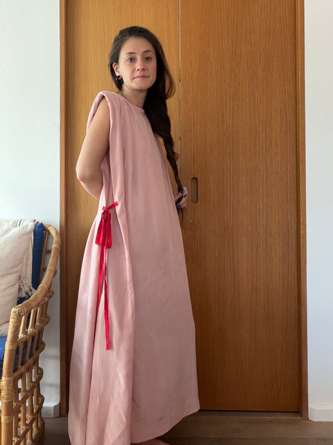 A woman wearing a long, sleeveless dusty pink dress or tunic with bright red tie details at the waist, standing in front of a wooden door in an indoor setting.
