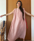 A woman wearing a sleeveless, loose-fitting dusty pink maxi dress with a red tie detail at the side, standing in front of a wooden door.