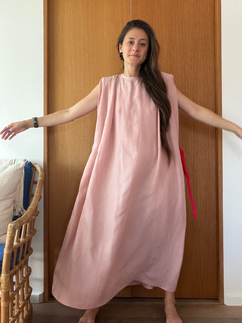 A woman wearing a sleeveless, loose-fitting dusty pink maxi dress with a red tie detail at the side, standing in front of a wooden door.