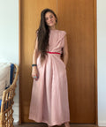 A woman stands barefoot in front of a wooden door wearing a dusty pink midi dress with a thin red waist tie. A wicker chair is partially visible on the left.