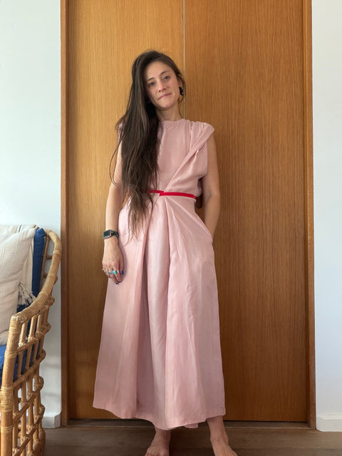 A woman stands barefoot in front of a wooden door wearing a dusty pink midi dress with a thin red waist tie. A wicker chair is partially visible on the left.