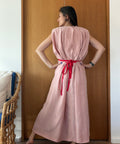 A woman stands with her back to the camera in front of a wooden door, wearing a sleeveless dusty pink maxi dress with a bright red tie belt at the waist.