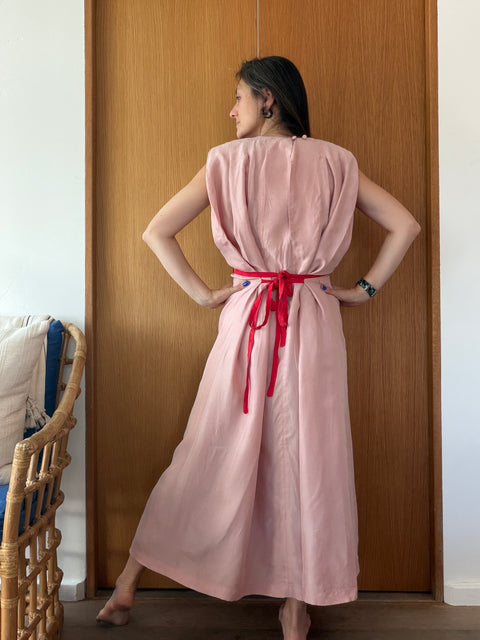 A woman stands with her back to the camera in front of a wooden door, wearing a sleeveless dusty pink maxi dress with a bright red tie belt at the waist.